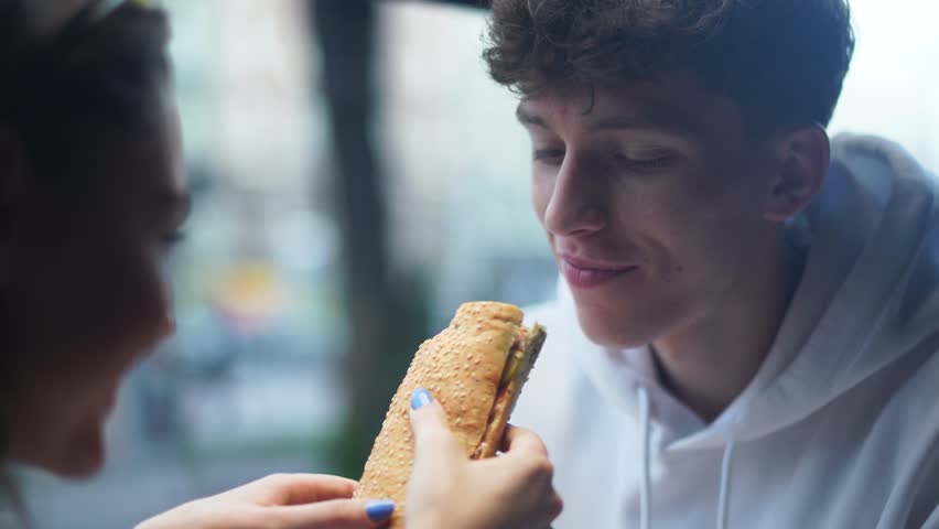 Girl cheerfully and playfully feeds her smiling boyfriend with sandwich and then gently wipes his mouth. Young couple in the fast food store.