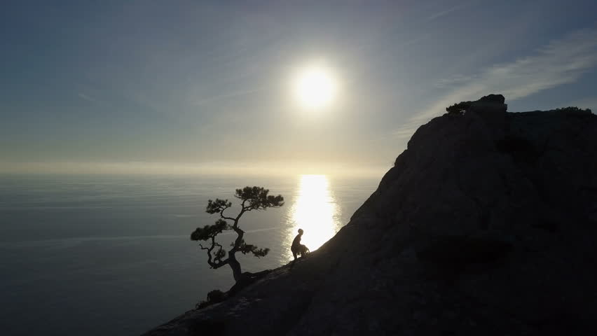Flight over of young woman standing on the top of a mountain facing the sea. Lady on the summit in beautiful scenery.