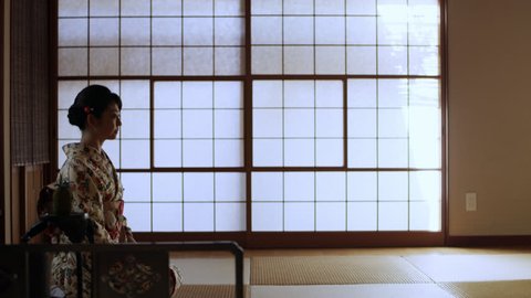 Woman profile peacefully sitting in a room being served tea at tea ceremony in a traditional Japanese home by man with soft day lighting. Medium shot on 4k RED camera on gimbal.