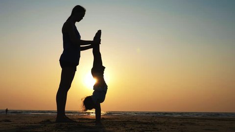 Mom train little girl doing handstand position. Silhouette of young sporty mother with little daughter doing gymnastic on the beach at sunset. Acrobatic exercise outdoor.