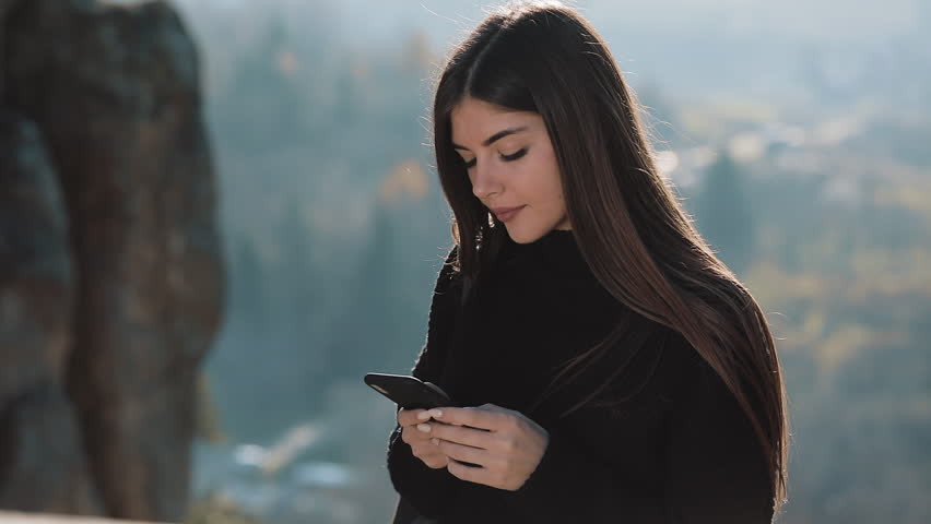 Young happy woman chats with someone on his phone standing on the top of the rock before beautiful mountain. Communication, tourism, hike concept