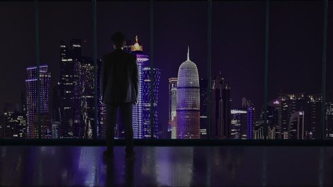 Silhouette of the businessman in the office standing in front of a big window with a view of Doha, Quatar. Folding arms.