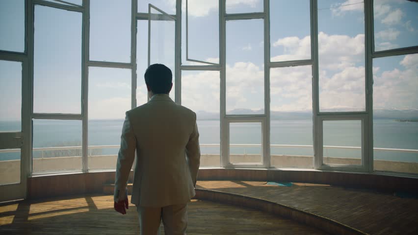 Businessman in a suit is walking towards a big window that looks at the sea . Back View of the Businessman wearing a Suit Standing in empty place . Shot on ARRI ALEXA cinema camera in slow motion . | Shutterstock HD Video #1022205076