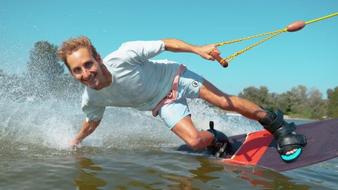SLOW MOTION CLOSE UP Cheerful young surfer wakeboarding, sliding his hand on water surface, splashing water at camera. Smiling wakesurfer riding waterski cable park on sunny day. Wakesurfing in summer