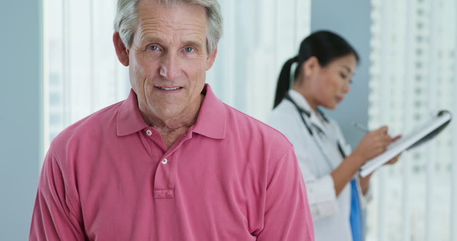 Portrait of senior Caucasian male patient looking at camera and smiling while doctor works in background. Attractive older man in hospital with female physician out of focus behind him. Slow motion 4k | Shutterstock HD Video #1022234977