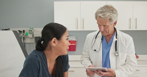 Older man doctor listening to medical history from Japanese woman in exam room while holding tablet computer. Senior Caucasian male medical professional in OBGYN office with patient. Slow motion 4k