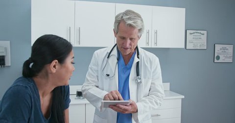 Older man doctor taking medical history on tablet computer from Japanese woman in exam room. Senior Caucasian male medical professional in OBGYN office with patient. Slow motion 4k