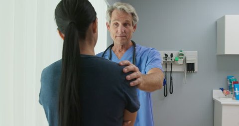 Medium shot of supportive senior Caucasian male doctor listening to female patient with his hand on her shoulder in hospital exam room. Woman talking to her nurse or OBGYN. Slow Motion 4k