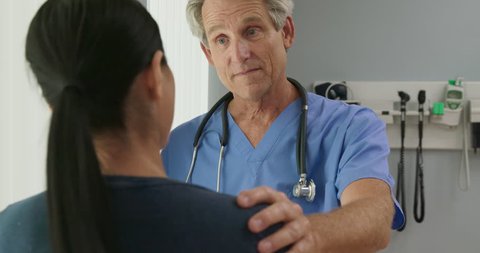 Over the shoulder shot of supportive senior Caucasian male doctor listening to female patient with his hand on her shoulder in hospital exam room. Woman talking to her nurse or OBGYN. Slow Motion 4k
