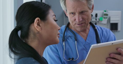 Over the shoulder shot of doctor showing tablet computer with results to patient in hospital exam room. Senior Caucasian male medical professional and Asian woman going over records. Slow Motion 4k