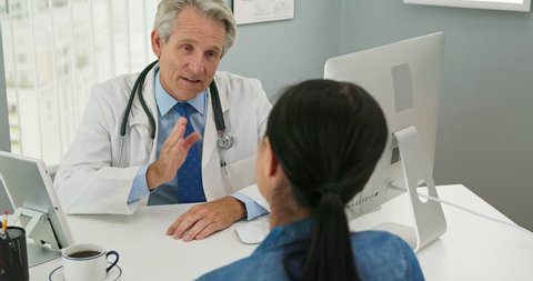 High angle view of senior Caucasian male doctor in his office talking to new patient and smiling. Primary care physician explaining medical issue to woman as camera cranes down. Slow motion 4k