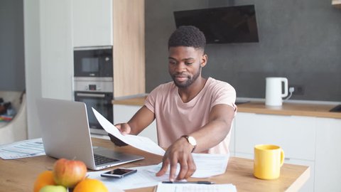 Handsome dark-skinned man having thoughtful and serious facial expression while reviewing finances and paying on-line bills, sitting at desk with documents in modern home interior