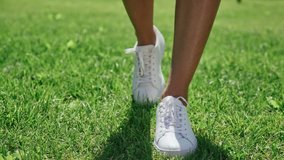 Female feet in white sneakers, are walking along the bright green grass. Walking on the lawn in white shoes: close-up, only legs. A shadow on the grass. Slender female legs in white shoes