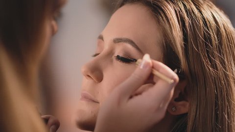The make-up artist gently puts a black eyeliner on the eyelids of the eyes, with a professional brush on the face of a Caucasian blonde model. Profile view. Close-up, make-up business woman. Slow