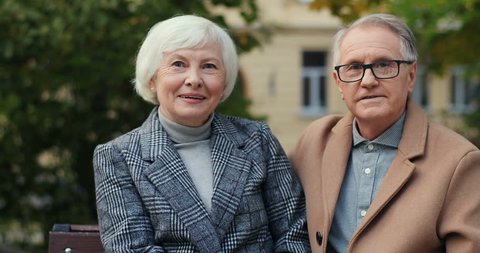 Portrait of the Caucasian senior couple sitting on the bench in autumn and smiling to the camera. Outdoor.
