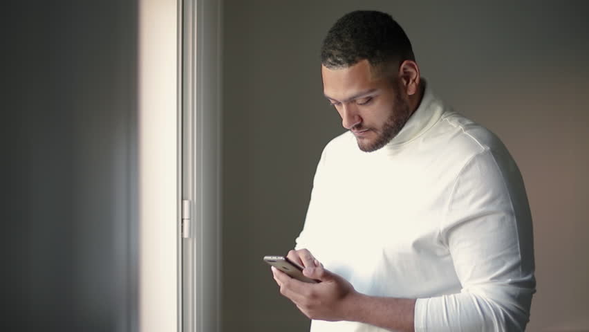 Pensive mix raced guy browsing news indoors. Young man in white casual shirt using smartphone, sliding finger across screen. Mobile internet concept
