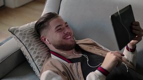 Cheerful guy using tablet for video call at home. Young Caucasian man in earphones lying on couch, holding tablet and waving hand to screen. Video call concept