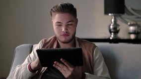 Serious stylish guy using tablet, becoming cheerful and positive. Young Caucasian browsing web at home and getting good news. Good news concept