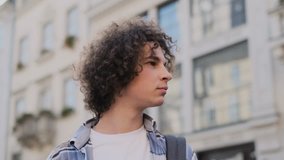 Close up portrait of happy cute caucasian man smiling in city. Handsome young curly man, tourist or student, millennial in hipster outfit, outdoor. Trendy man looking at camera