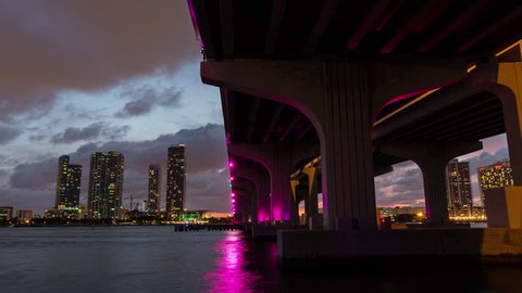 Day to night sunset timelapse hyperlapse view I-195 Highway Bridge in Miami. Concrete pillar and skyscraper buildings. Light reflection in the bay water.