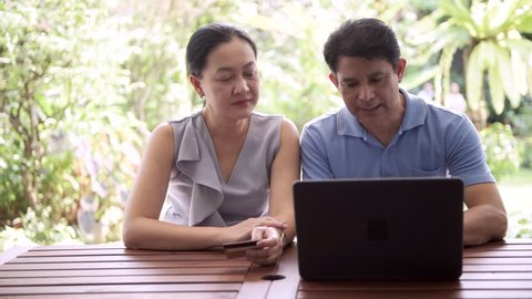 Senior asian man trying to key in credit card number that his wife is holding into laptop outdoor with beautiful tropical garden in background. Senior online shopping concept.