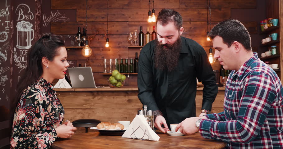 Bearded waiter bringing cofffee and croissants to a gorgeous inlove couple. They are in a rusting coffee shop with wooden walls