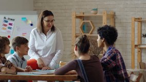 Tracking medium shot of middle-aged woman in glasses sitting on desk and talking to multi ethnic group of four teenagers during workshop - Powered by Shutterstock - Get 15% off with code: PIKWIZARD15