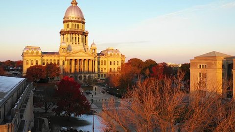 Stunning rising slow motion aerial drone shot of the Illinois State Capitol Building in Springfield, Illinois, at dawn in November as the sun rises and fall leaves glow orange in the distance.