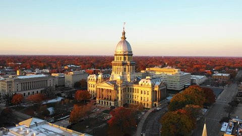 Stunning establishing aerial drone shot of the Illinois State Capitol Building in Springfield, Illinois, at dawn in November as the sun rises and fall leaves glow orange in the distance.