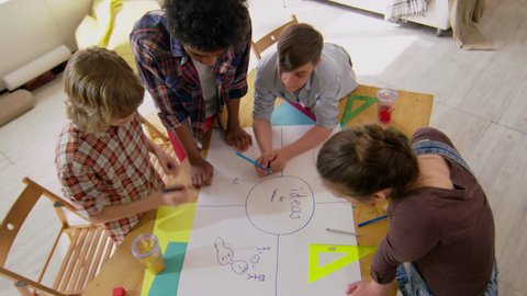 High angle view of group of four teenage students of different ethnicities leaning on desk and drawing mind map on paper together using colored pencils and felt tip pens