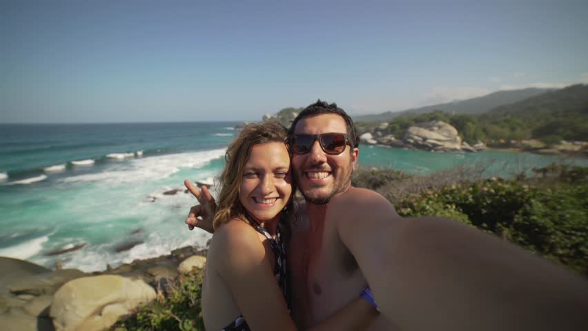 Happy couple taking selfie photo in front of the sea in Tayrona National Park, Tropical Colombia. Crazy tourists travelling on the white beach of caribbean sea. 4K video