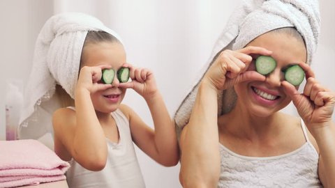 Wellness, spa, skincare concept. Young woman with daughter putting cucumbers on eyes, having fun after bath.  
