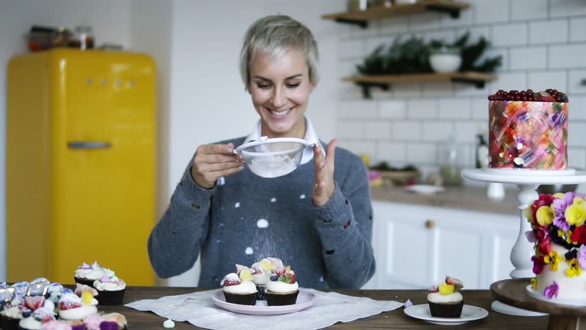 Young female confectioner decorating tasty, fancy decorated cupcakes. Woman with grey colored hair icing sugar sprinkles on cakes with fresh strawberries and cream on the top. Front view. Slow motion