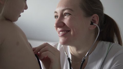 Beautiful doctor listens to the heartbeat of the child. A woman in a white coat, puts a stethoscope to the breast of a child, smiles and listens to a little girl. A child in a hospital ward. Close-up
