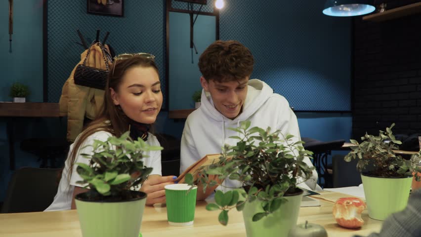 Girl reads her boyfriend a funny excerpt from fiction book and they laugh and smile sitting in coffee house