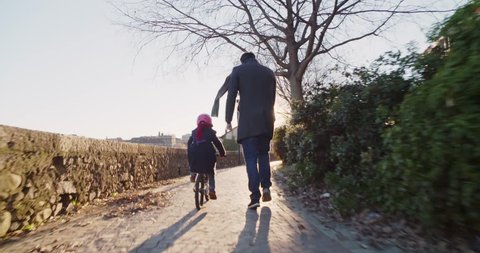 Daughter child girl learning riding bycicle with dad teaching in city.Growing,childhood,active safety family.Sidewalk urban outdoor.Warm sunset cold weather backlight.4k slow motion 60p back video