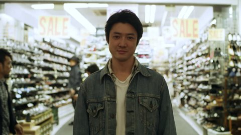 Portrait of a content Japanese man standing in front of a brightly lit store with people walking behind him on the street at nighttime. Medium shot on 4k RED camera.