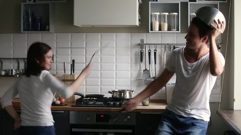 Funny young couple pretending fight battle kitchen war holding utensils having fun cooking together, happy playful man and woman laughing enjoying preparing food struggling with kitchenware at home