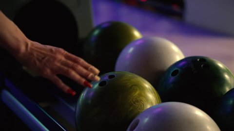 A girl chooses a ball before throwing while playing bowling in a bowling club. Beautiful manicured woman’s hands with red nail polish. A girl holding a bowling ball. Closeup. Sports and entertainment