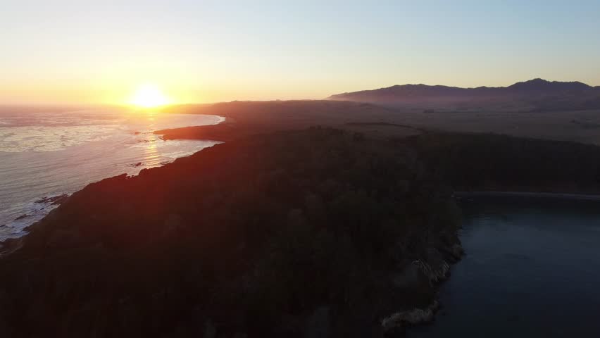 Aerial pull back shot of partial silhouette coastline over sea waves at sunset.