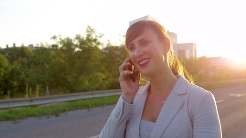 SLOW MOTION CLOSE UP: Stylish businesswoman talking on her phone while walking down sidewalk at sunset. Cheerful young woman in a grey suit calling her friends after a long day at work in the city.