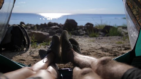 Close-up view of young couple's legs lying in camping tent with feet joined while resting after hiking day. Happy lovers enjoying beautiful sea view and relaxing in the evening.
