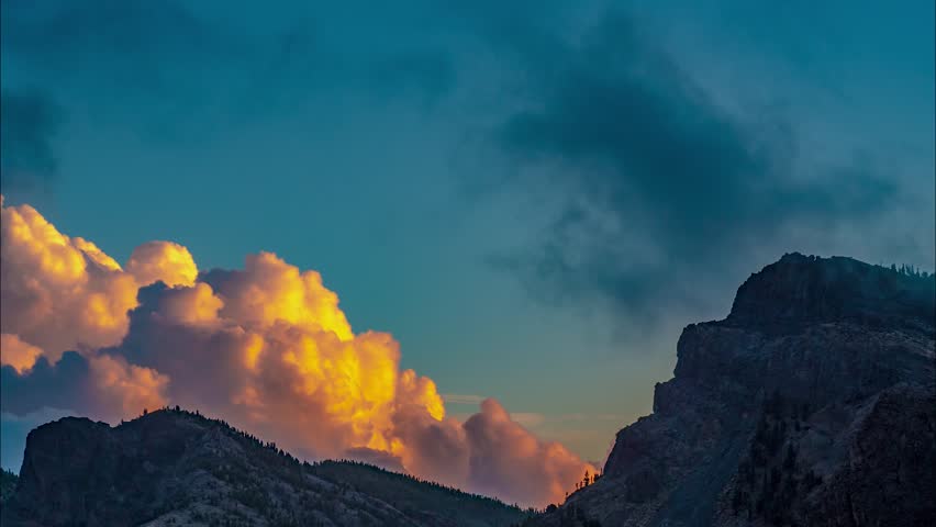 Sunset light shines on tall dramatic clouds behind two mountain peaks in Tenerife. Jagged cliff faces in shadow as fog and clouds boil around them in time lapse view. TILT DOWN toward tree line.