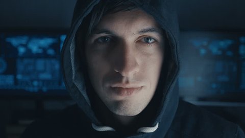 Close up Portrait of Hacker programmer in Black Hoody at background of cyber security center filled with display screens.