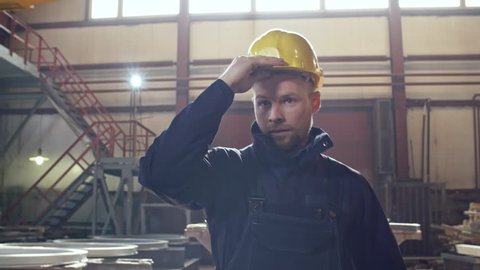 Dolly shot of tired worker in overalls walking through metal fabrication facility of steel plant and putting on hard hat and gloves