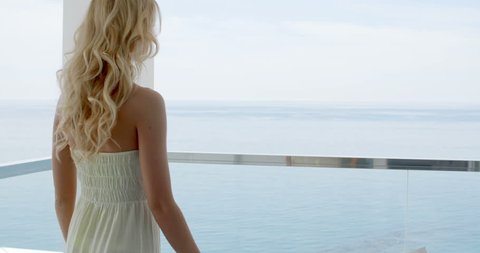 Rear View of Blond Woman Wearing Long Strapless Summer Dress Leaning on Railing of Glass Balcony Overlooking Ocean