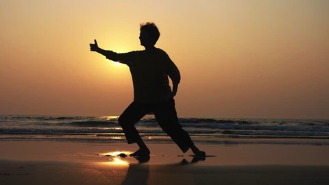 Silhouette of active senior woman practicing tai chi gymnastic on sandy beach at sunset in slow motion