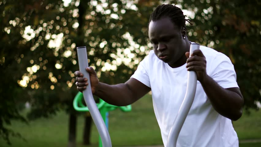 African man working out rigorously on a cross trainer, at a outdoor gym in a park at golden hour