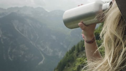 Blonde girl with hat drinking water from stainless steel bottle while enjoying the mountain view of slovenian alps, over shoulder