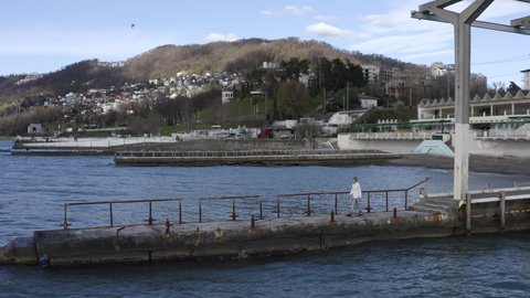 Man walking on pier outstretching arms and enjoying beautiful landscape drone view. Young man standing on sea pier and looking up. Embankment of marine city, building and highlands background